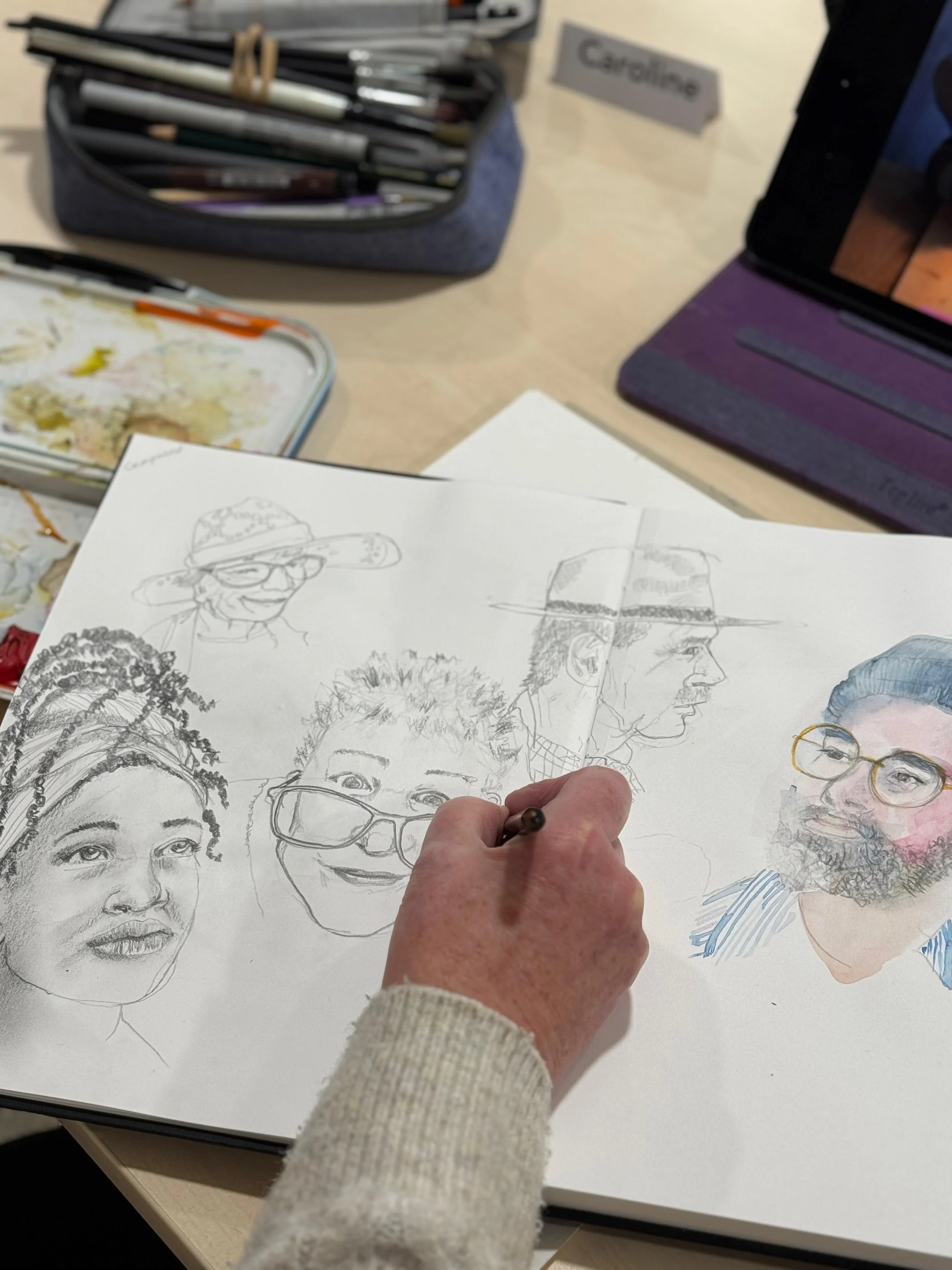 Women sitting at a long table with art supplies, painting and drawing during a watercolor workshop; large windows in the background let in natural light.