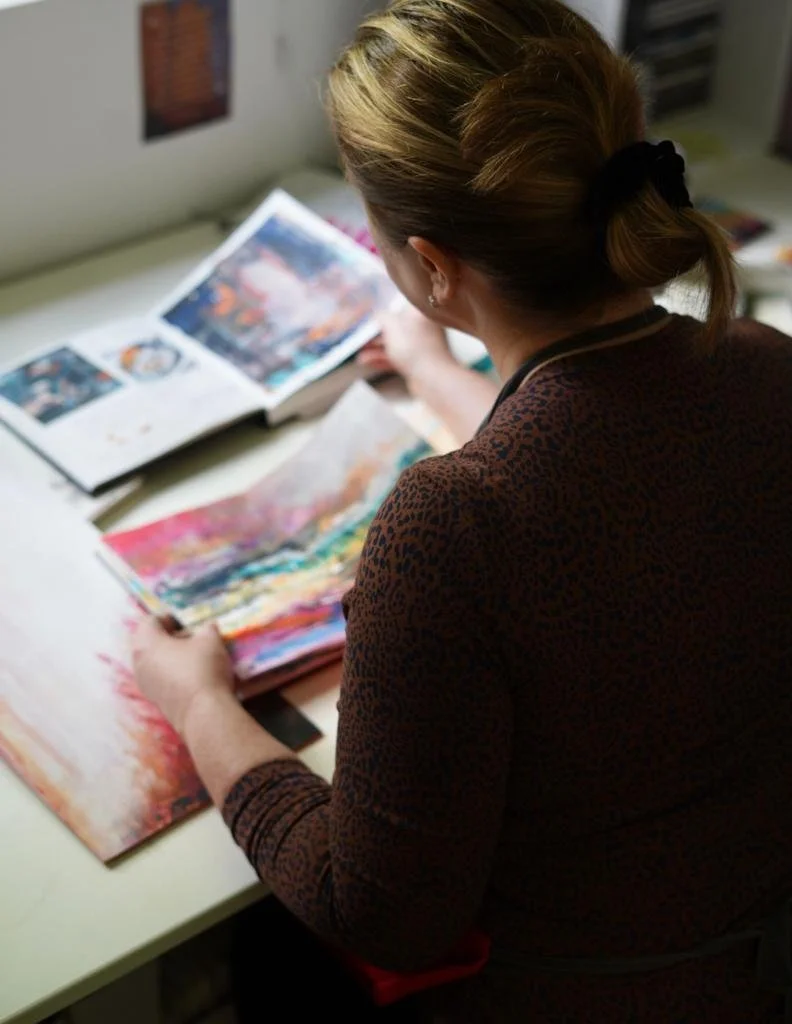 Jo Watson sitting at a table, flipping through colorful art books with abstract paintings.