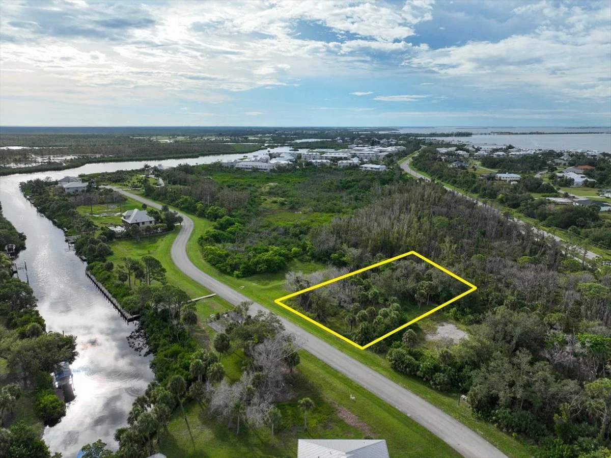 Aerial view of a lush, green residential area with lots of trees, waterways, and lots of houses. A small, empty lot outlined in yellow is in the foreground next to a gravel driveway and a road.