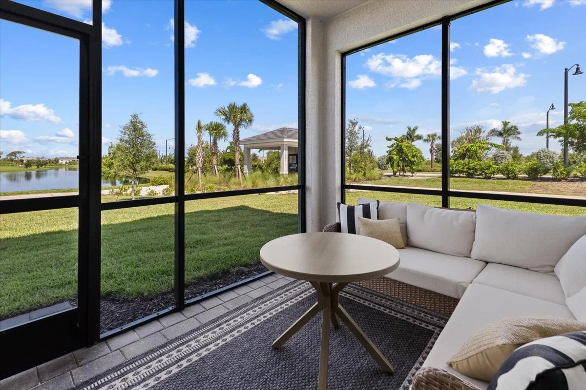Screened porch with outdoor seating, wicker sectional sofa with white cushions and pillows, round table, and views of a green lawn, trees, and sky.