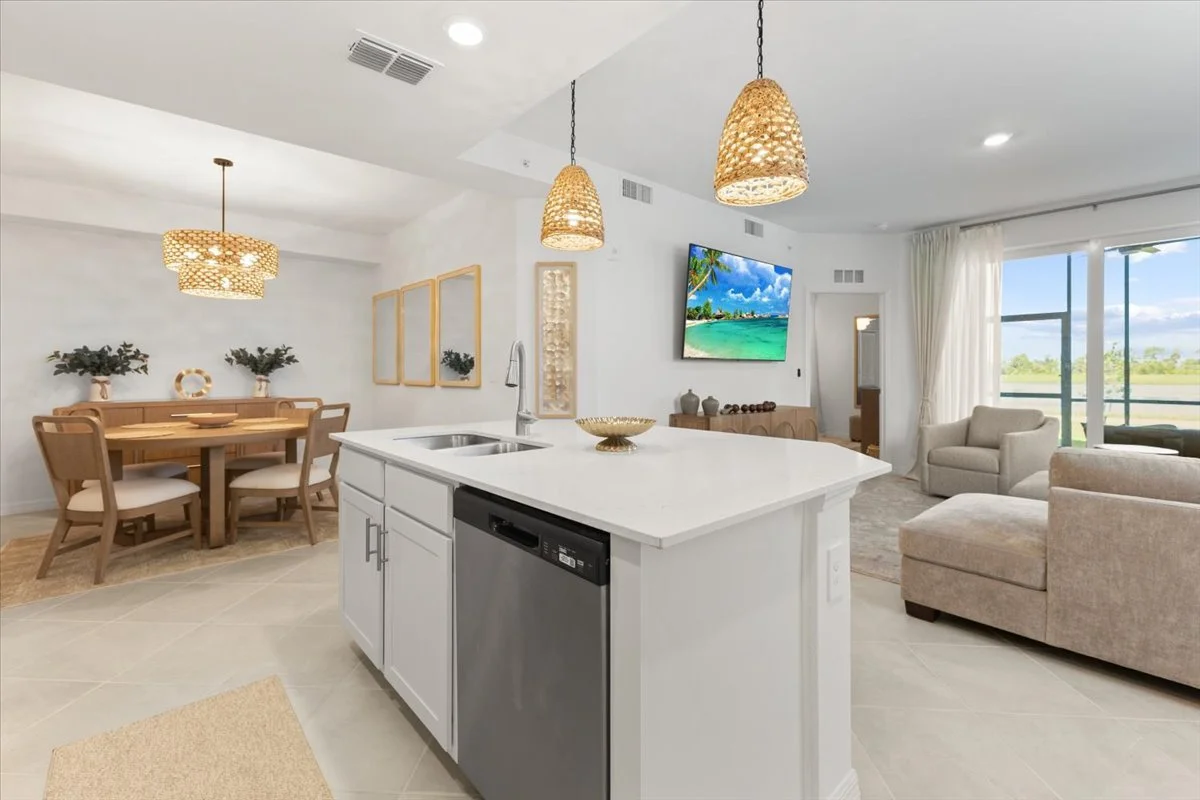 Open-concept living room and dining area with white walls, large windows, and a kitchen island with a sink and dishwasher.