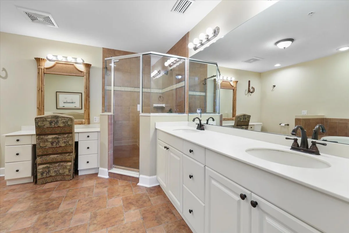 A spacious bathroom with double sinks, a large mirror, a glass-enclosed shower, a vanity with a chair, and warm-toned tile flooring and walls.