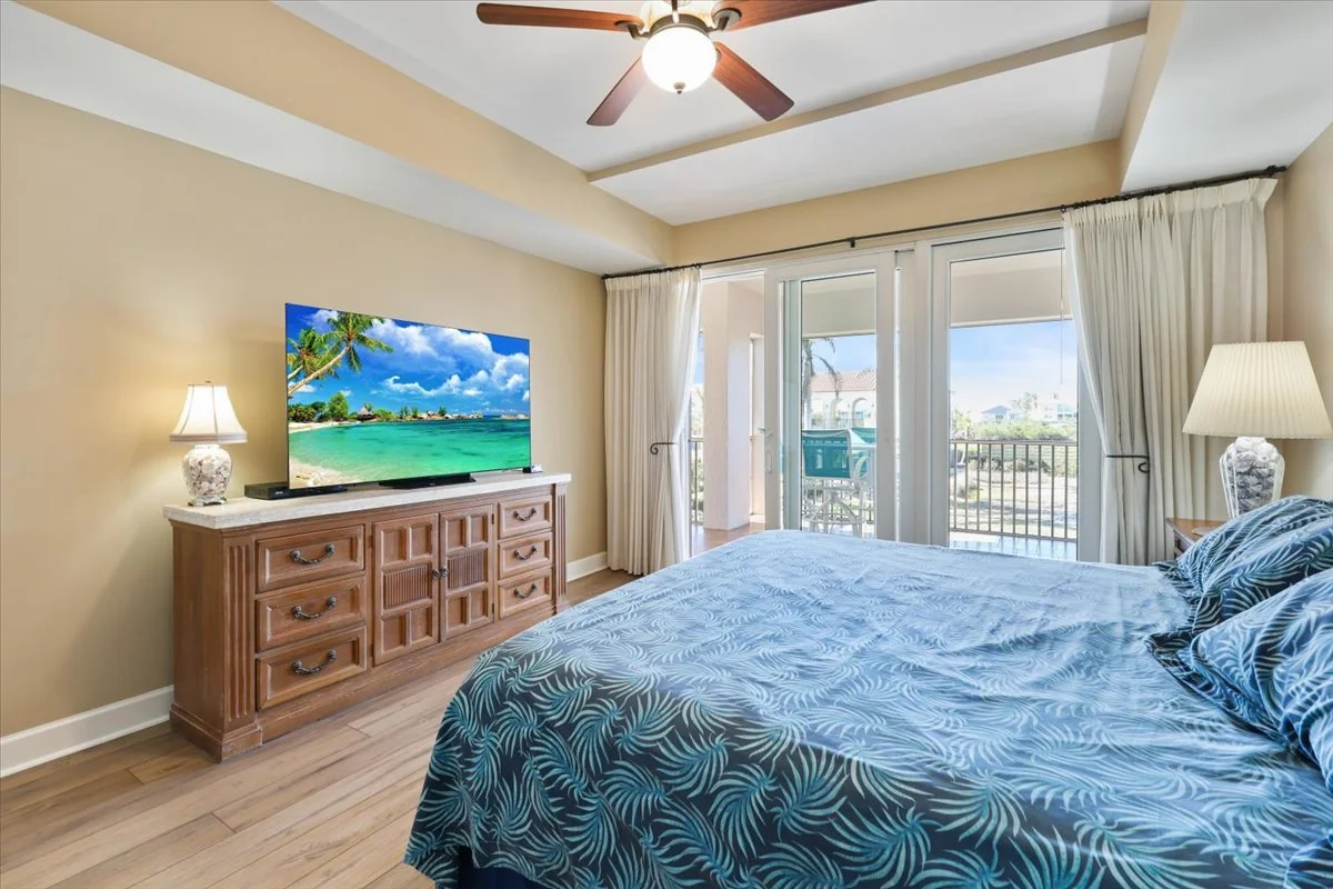 A bedroom with a large bed covered in a blue tropical-patterned bedspread, two lamps on bedside tables, a wooden dresser with a flat-screen TV displaying a tropical beach scene, and glass sliding doors opening to a balcony with outdoor furniture and