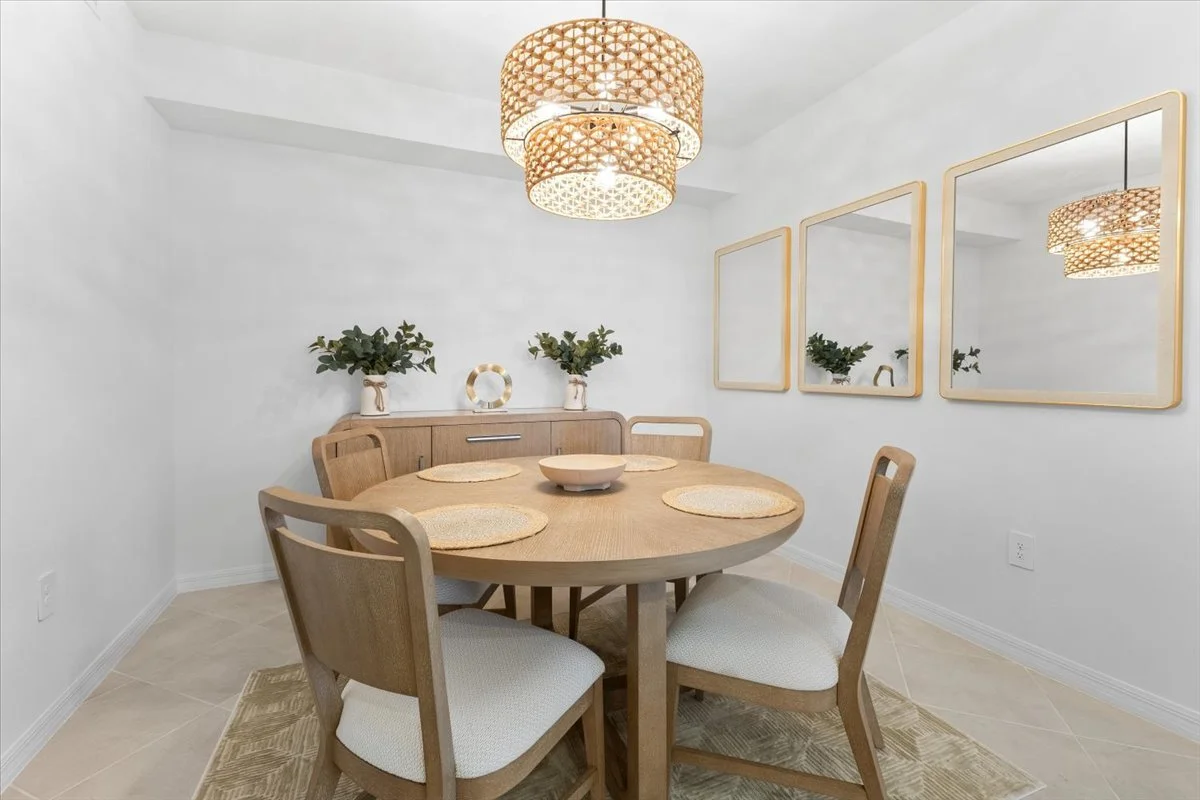 A minimalist dining room featuring a round wooden table with four matching chairs, a sideboard with potted plants and decor, three framed mirrors on the wall, and two woven pendant light fixtures.