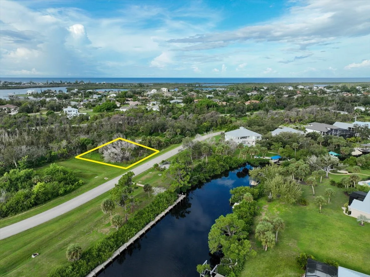 Aerial view of a residential area near water, with a vacant lot outlined in yellow, surrounded by trees and houses, near a canal with docks, and a distant ocean under a partly cloudy sky.