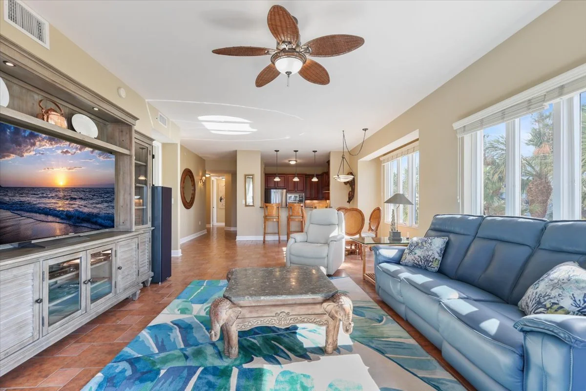 Living room with a large leather sofa, armchair, TV, coffee table, ceiling fan, and large windows showing palm trees outside.