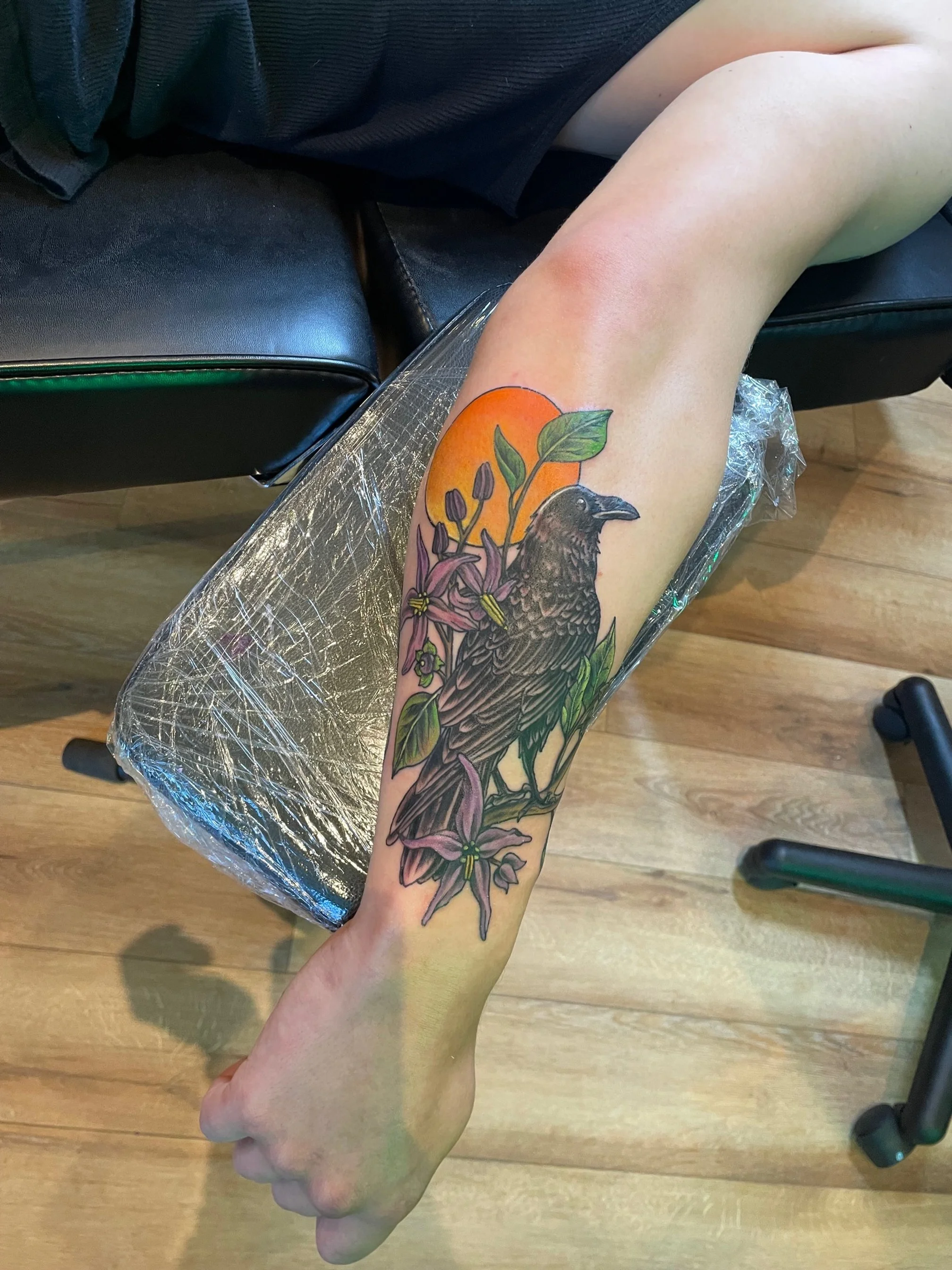Colorful tattoo of a bird perched on a branch with purple flowers, green leaves, and an orange sun in the background.