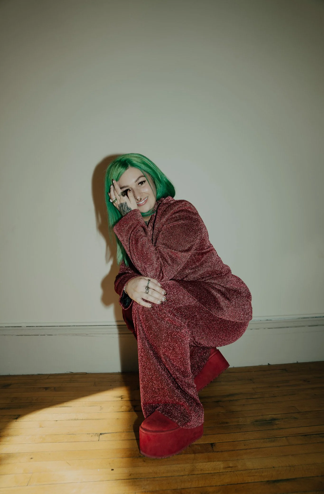 Person with green hair in a red outfit crouching indoors.