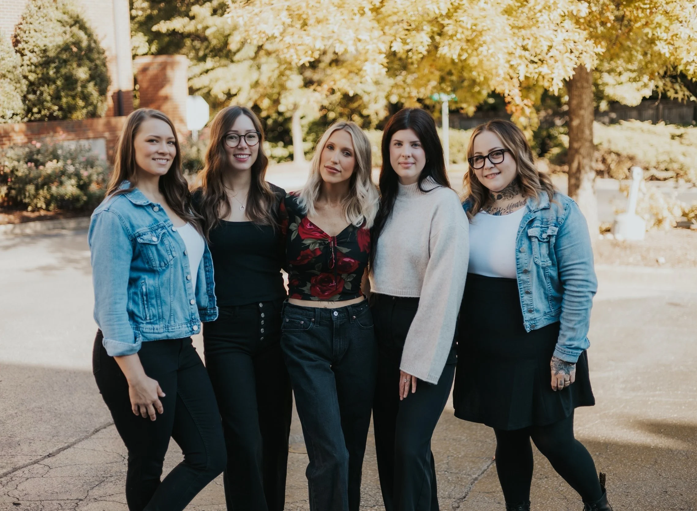 Five women standing outdoors, smiling, with trees in the background.