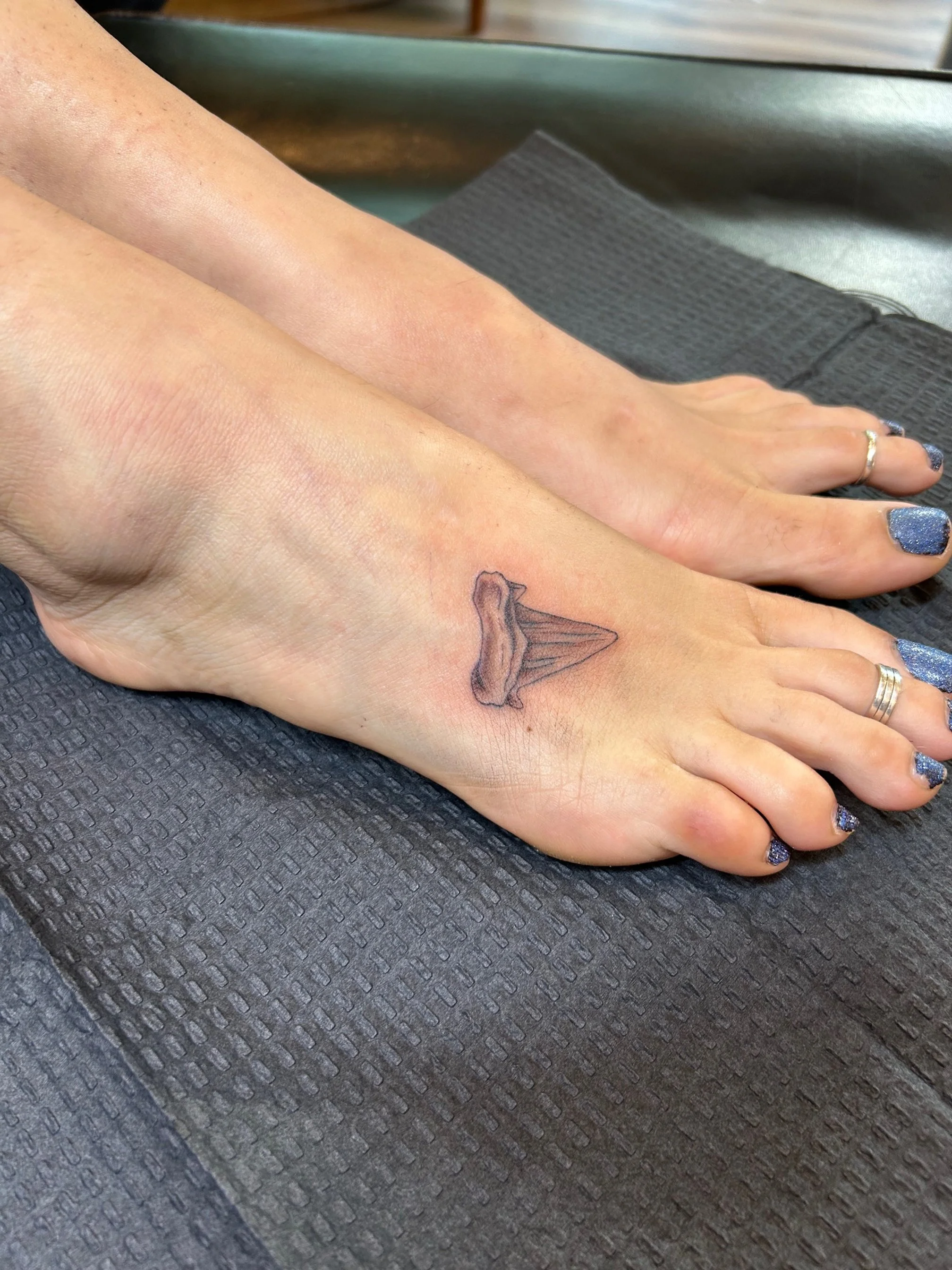 A person's foot resting on a black textured surface, with a small tattoo of an ice cream cone on the top of the foot near the ankle. The person has blue glitter nail polish on their toenails and are wearing rings on the toes.