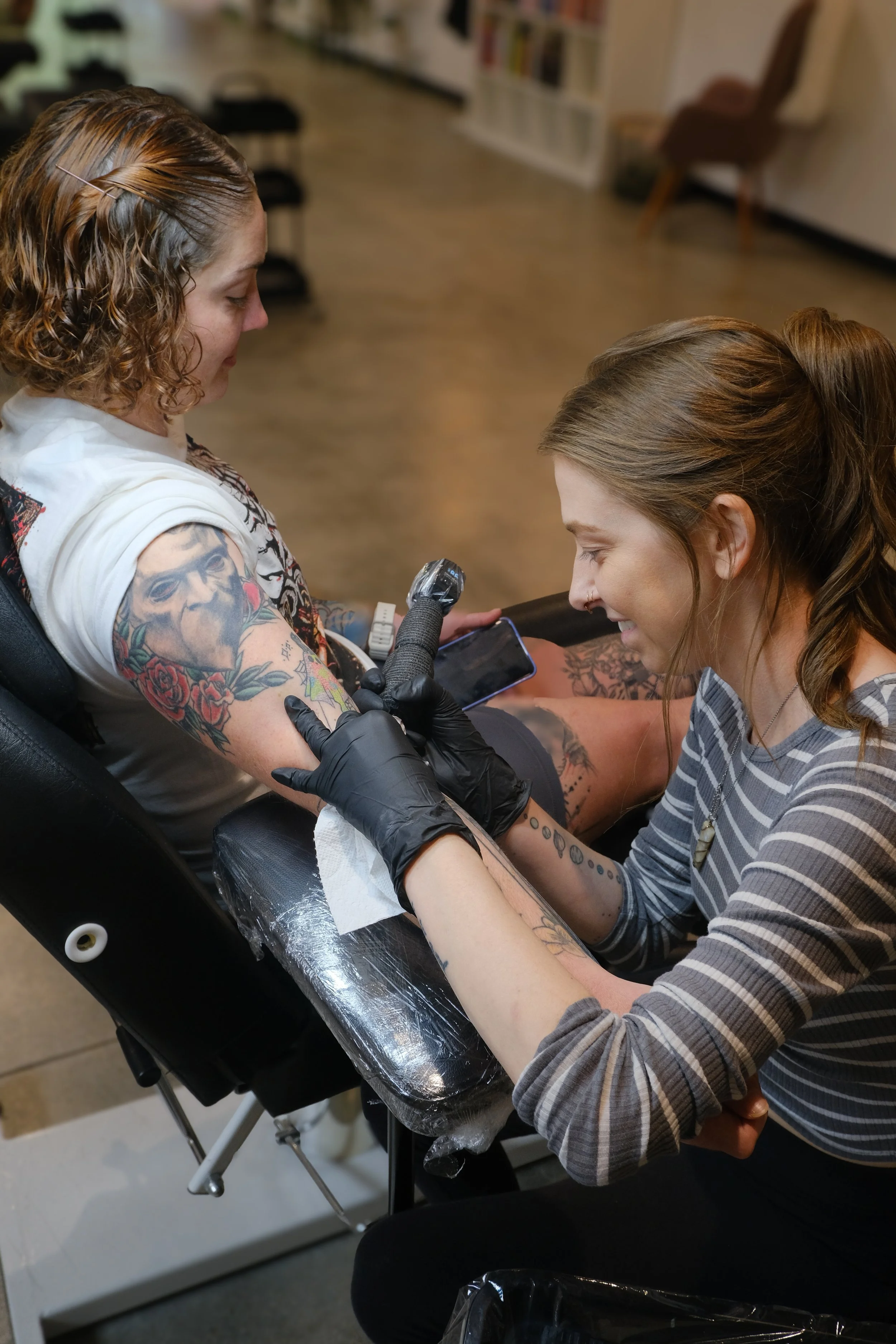 A woman with tattoos on her arm getting tattooed, while a tattoo artist with striped shirt and black gloves works on her arm in a tattoo parlor.