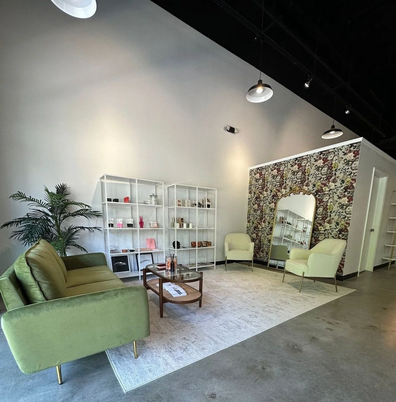 Modern waiting area with green sofa, white shelves displaying products, floral wallpapered mirror, and cream chairs.