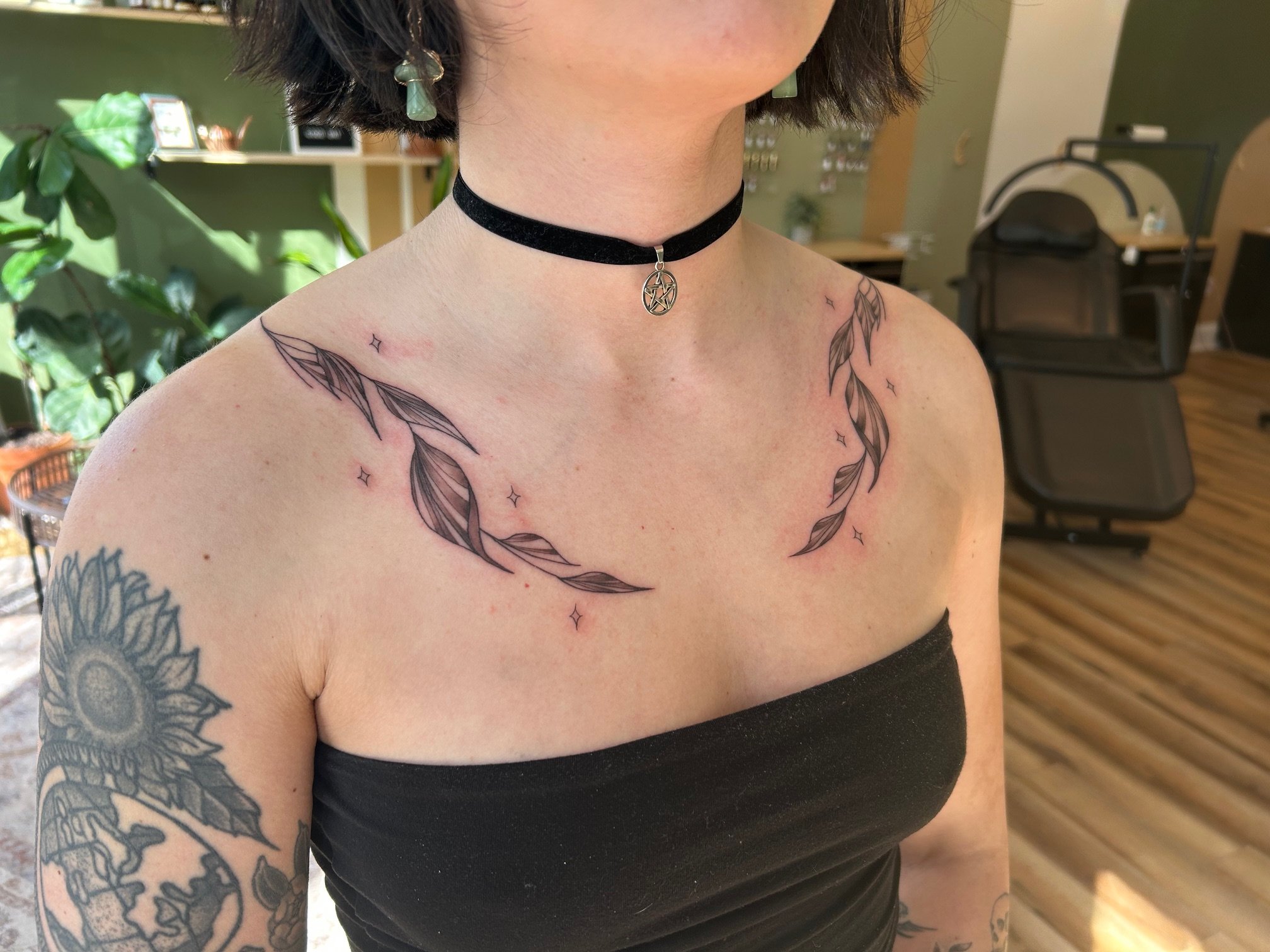 Person with short dark hair wearing a black choker with a pentagram pendant. They have tattoos on their shoulders and arm, including feathers on the shoulders and a sunflower on the arm. The person is wearing a black strapless top, and there is a gre