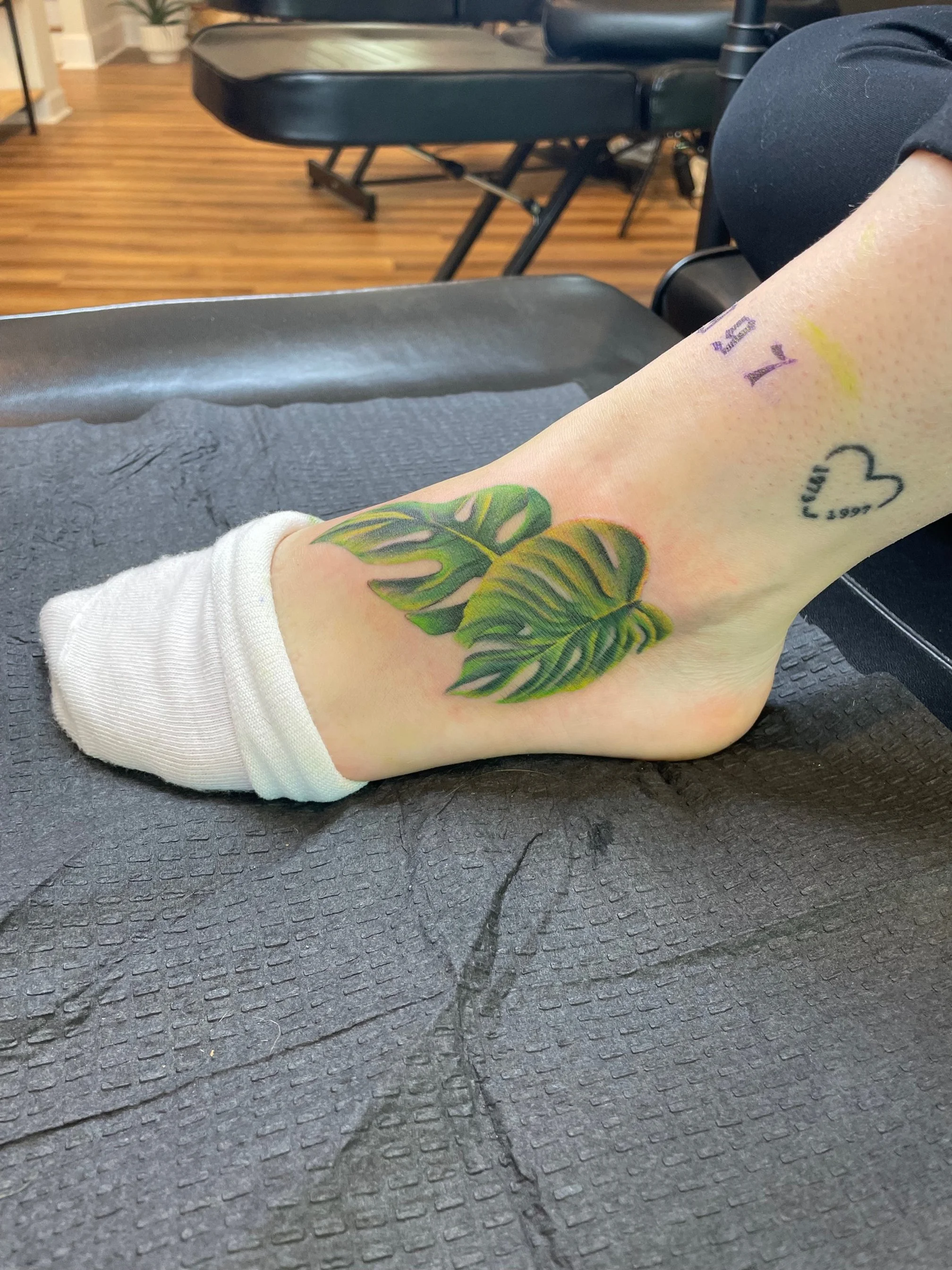 A tattoo of two green monstera leaves on a person's ankle, with other small tattoos nearby, one of a heart and one of the words 'I FELL.'