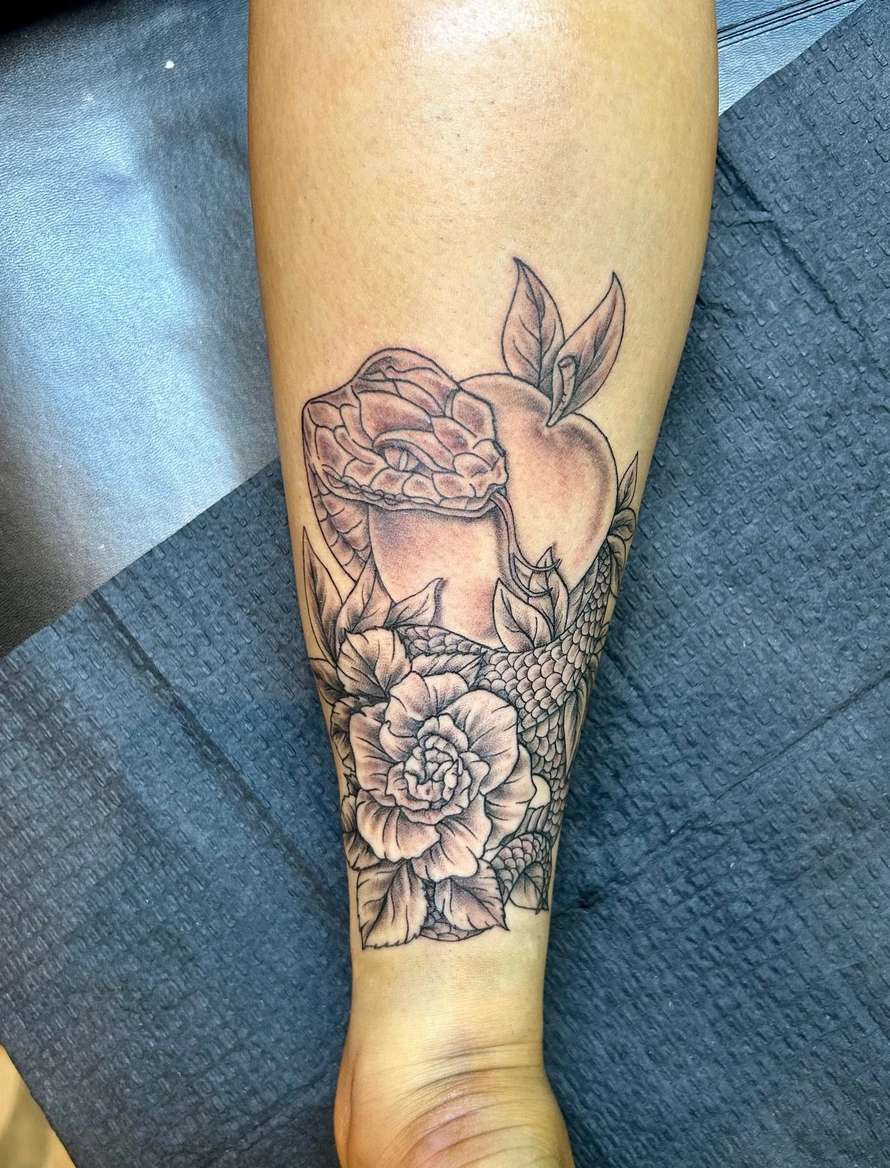 Black and gray tattoo on a person's forearm depicting a snake, a flower, and leaves with a peach in the background.