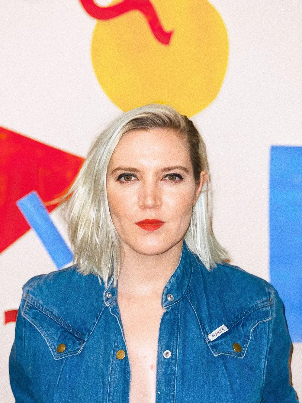 A woman with blonde hair and red lipstick posing in front of a colorful abstract wall.