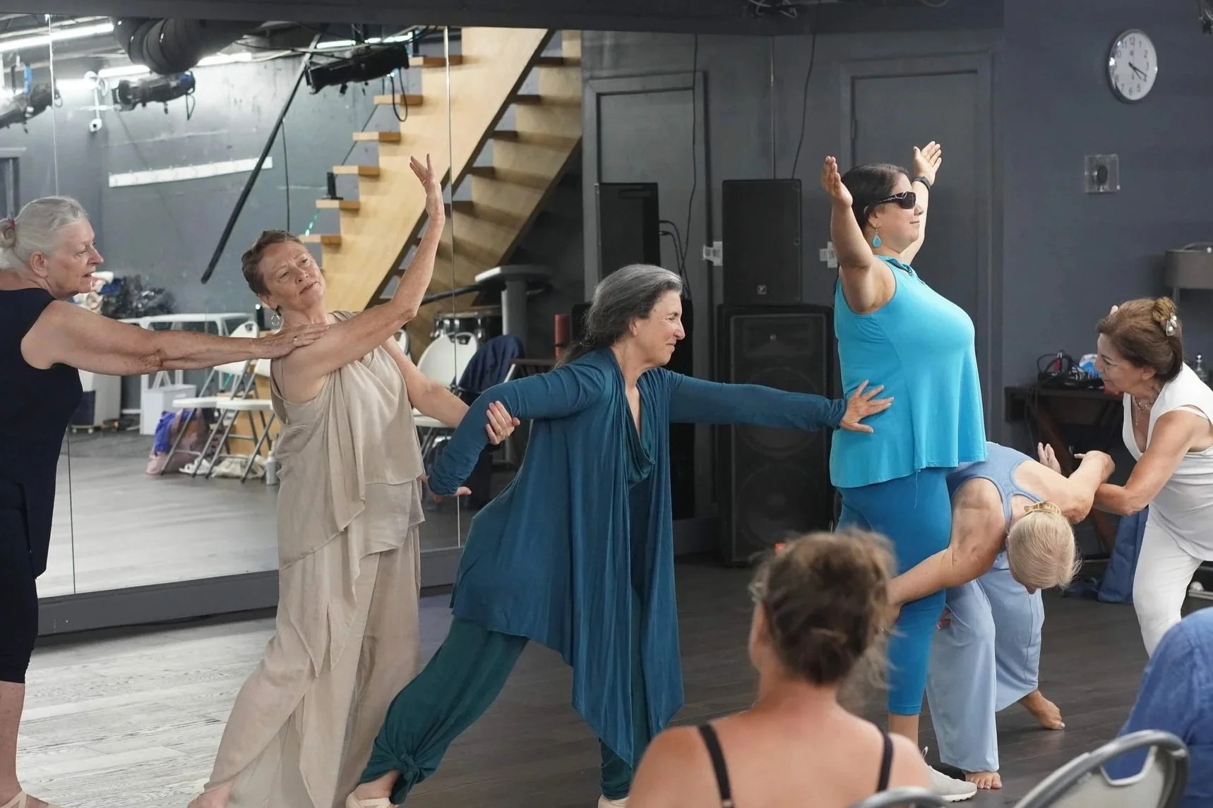 Group of older women participating in a dance presentation, with one woman in a blue top wearing dark glasses, in a studio with a mirror and various equipment.