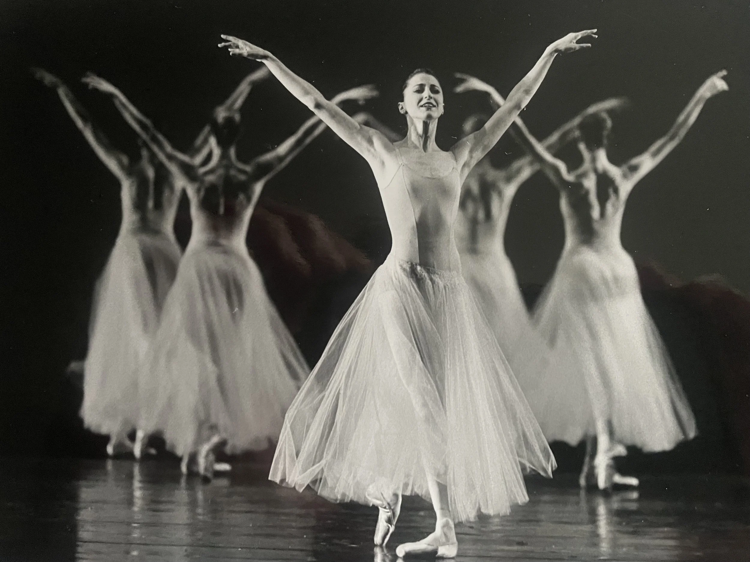 Black and white photo of ballerinas performing on stage, with one dancer in the foreground and others in the background, all wearing tutus and ballet shoes, with arms raised above shoulder height making a 'v'.