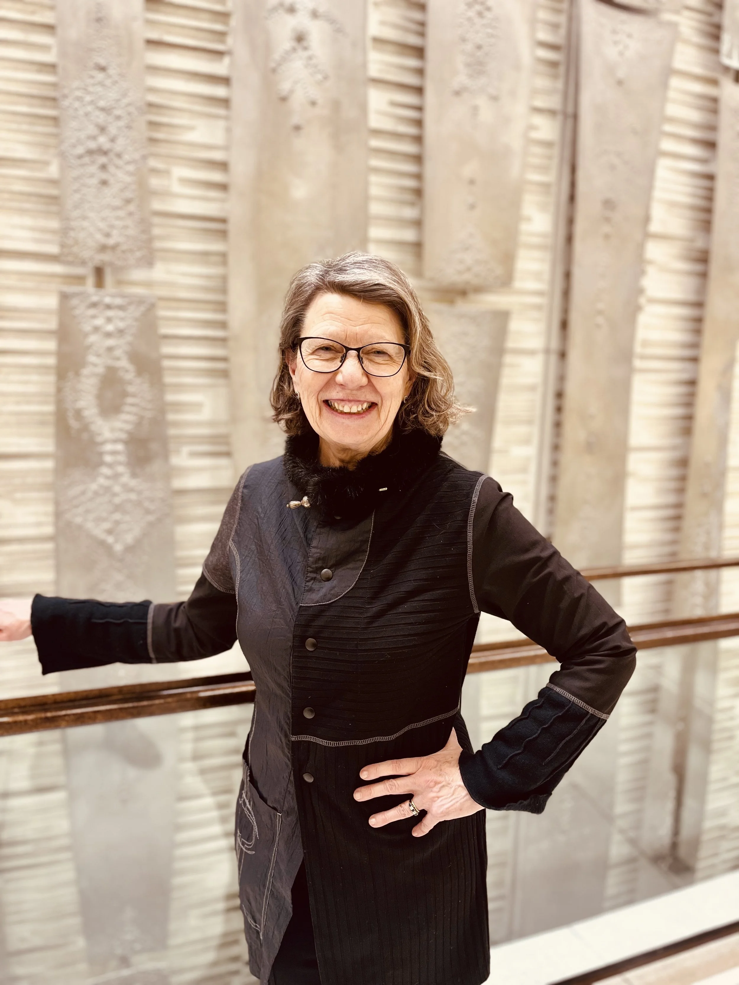 A smiling older woman with glasses, wearing a black jacket, standing indoors in front of textured beige wall art, with one hand on her hip and the other on a railing.