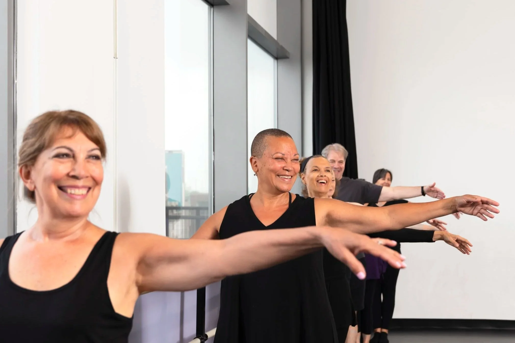 A group of diverse older adults in black dance clothing in a dance studio, smiling, with one arm extended to the side while other arm holds the ballet barre.