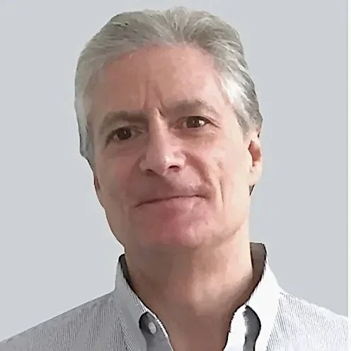 A person with a light gray hairstyle wearing a light-toned button-up shirt, looking at the camera with a neutral expression and a neutral background.