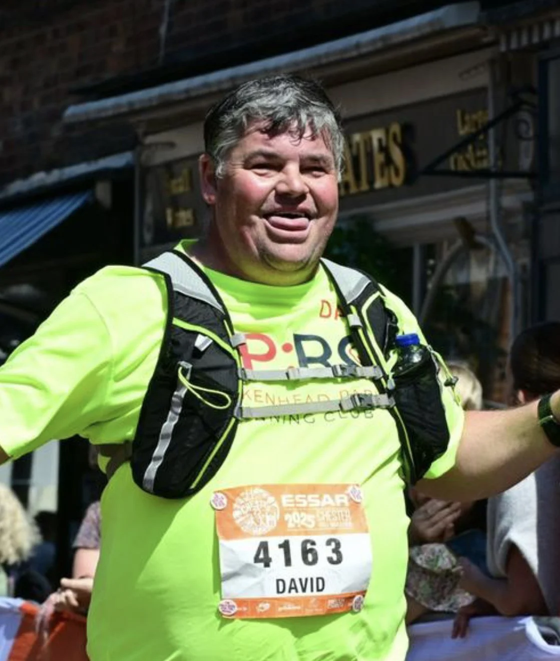A man wearing bright yellow running gear with a race bib reading '4163 DAVID' smiles and sticks out his tongue at a marathon or running event.