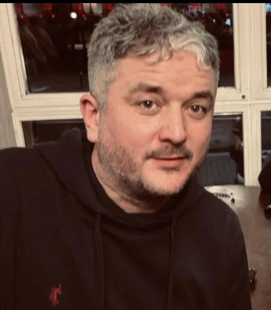 A man with short, light gray hair and a beard sitting in a room with large window panes behind him, wearing a black hoodie with a small red logo.