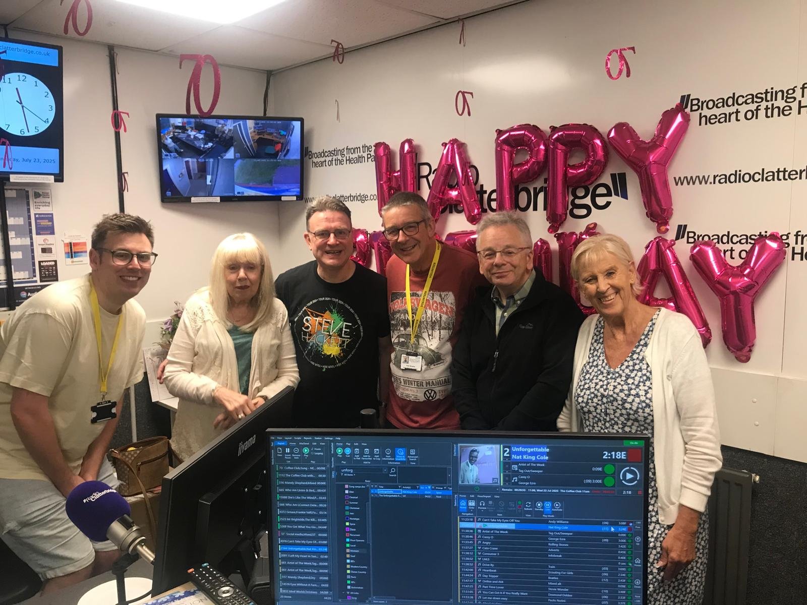 Group of six people posing and smiling in a radio station studio, with pink 'HAPPY' balloons and pink 'DAY' balloons on the wall behind them, broadcasting equipment visible in the foreground, and a digital clock and TV monitors on the wall.