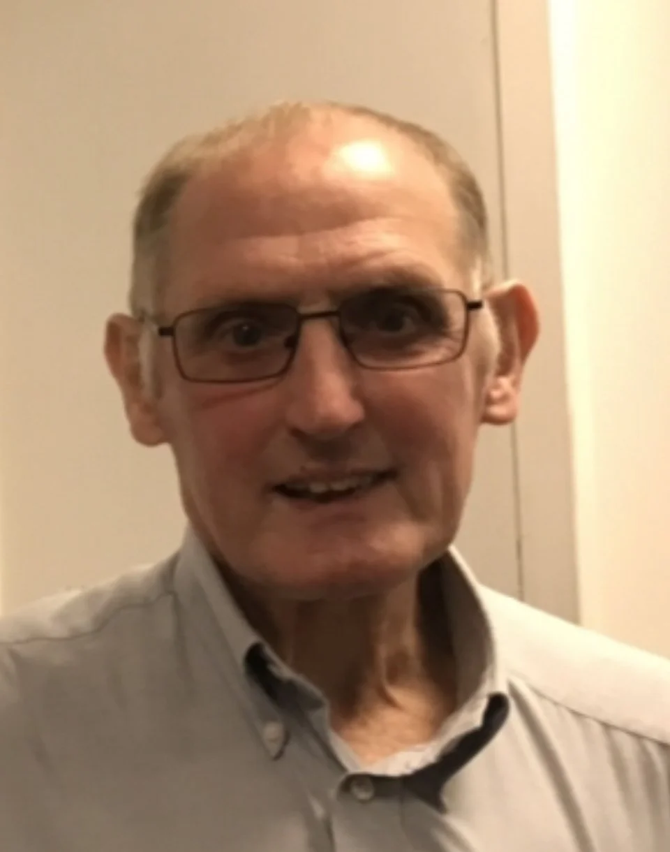 A middle-aged man with glasses, short light brown hair, and wearing a light gray collared shirt, standing indoors with a neutral background.