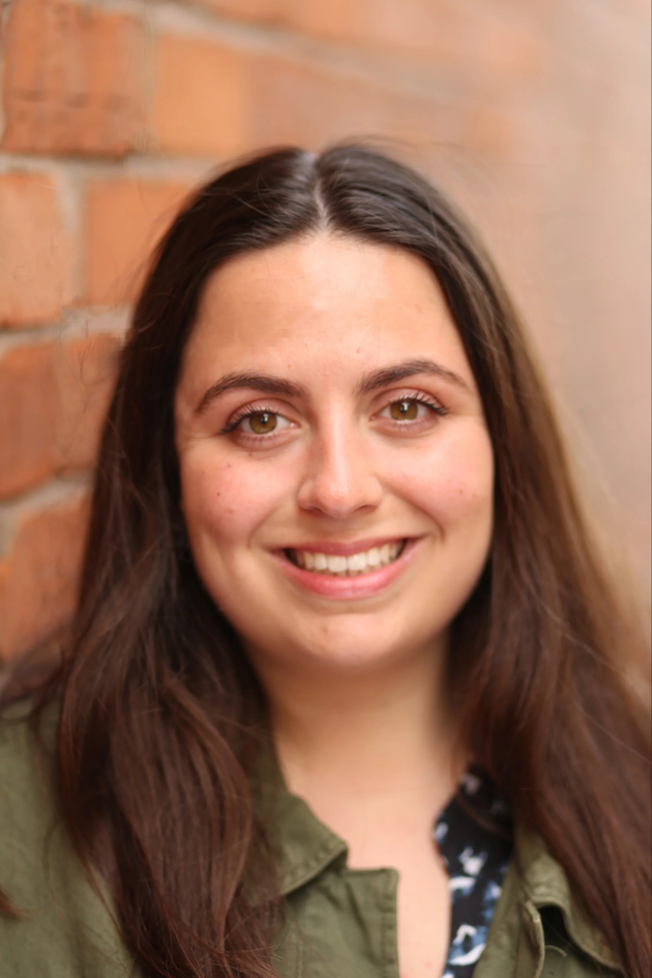 Close-up of a woman with long brown hair, smiling, with a brick wall background.