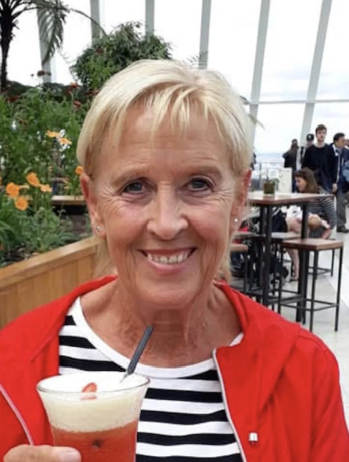 A smiling older woman with short blonde hair holding a layered cocktail drink with whipped cream and fruit, seated in a bright indoor space with large windows and plants.