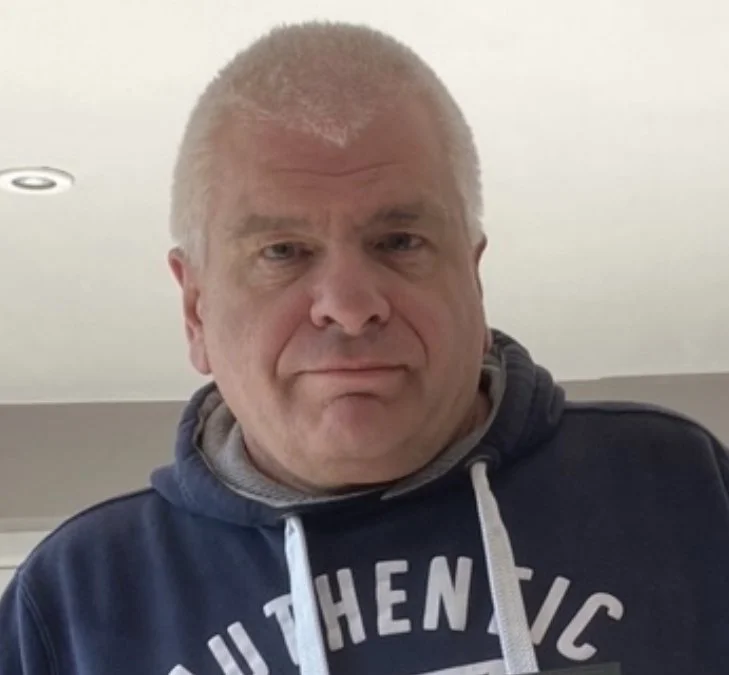 A middle-aged man with short, graying hair wearing a dark blue hoodie with white drawstrings and lens in the background.