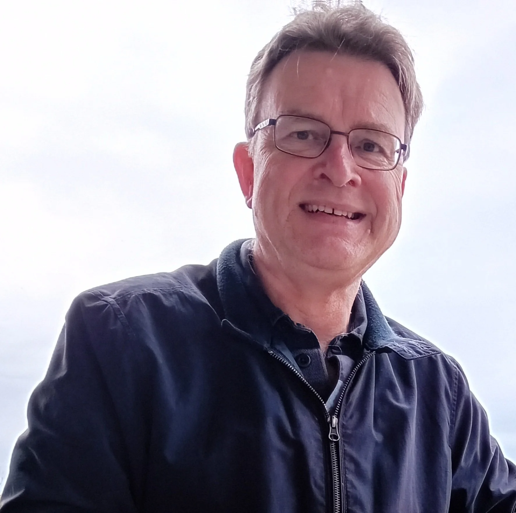 A middle-aged man smiling, wearing glasses, a dark jacket with a zipper, and a collared shirt underneath. The background is bright and plain.