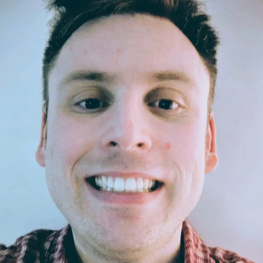 A close-up of a smiling young man with short dark hair, fair skin, and wearing a plaid shirt.