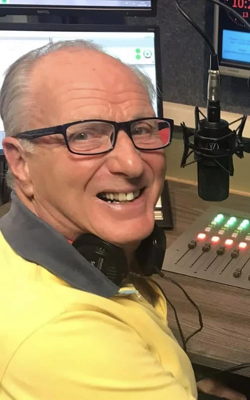 An older man with glasses smiling in a radio station studio, sitting at a soundboard with a microphone in front of him.