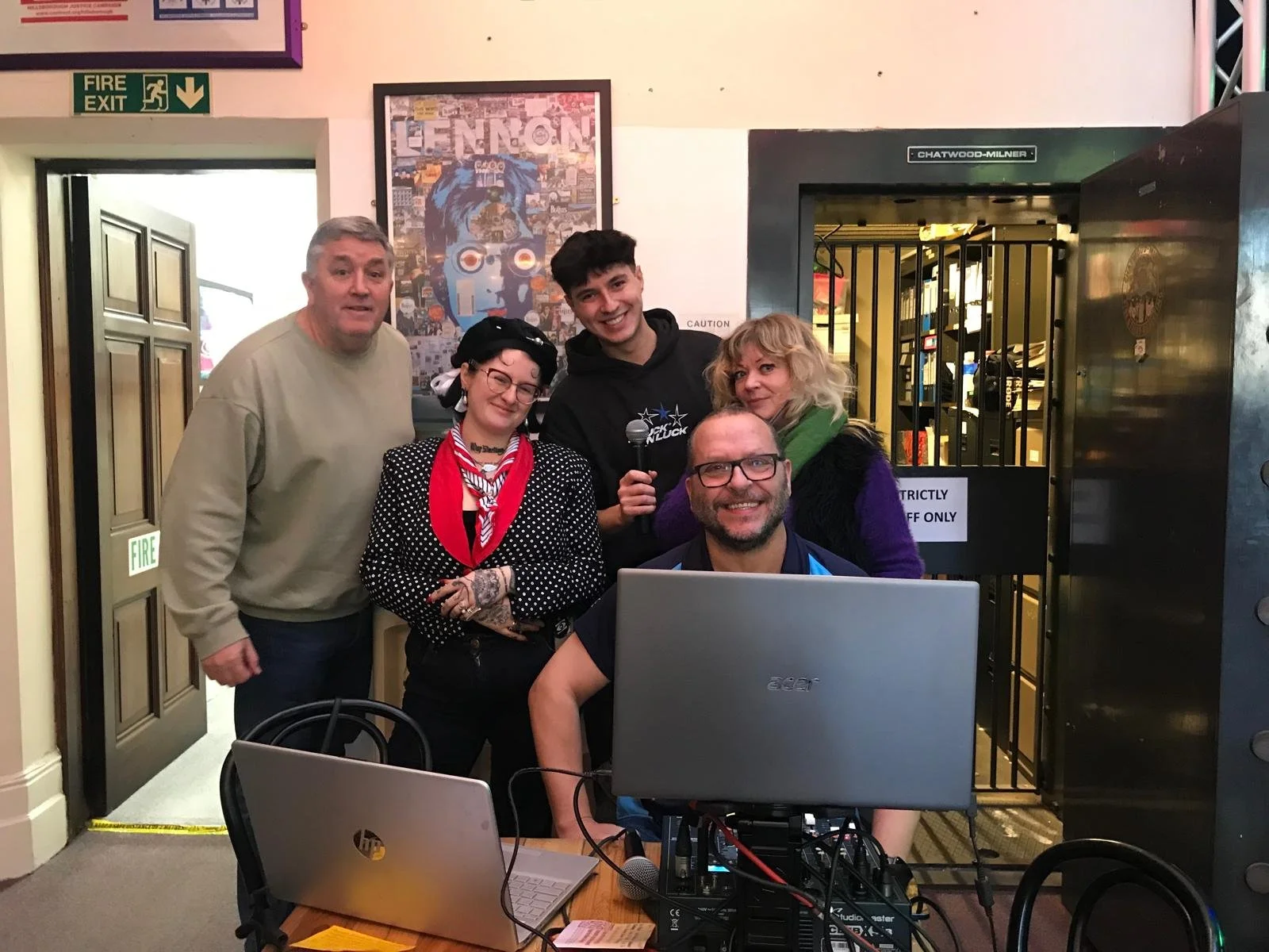 A group of five people standing behind a table with laptops, smiling at the camera in a room with a colorful poster and a door with a sign that says 'STRICTLY STAFF ONLY'.