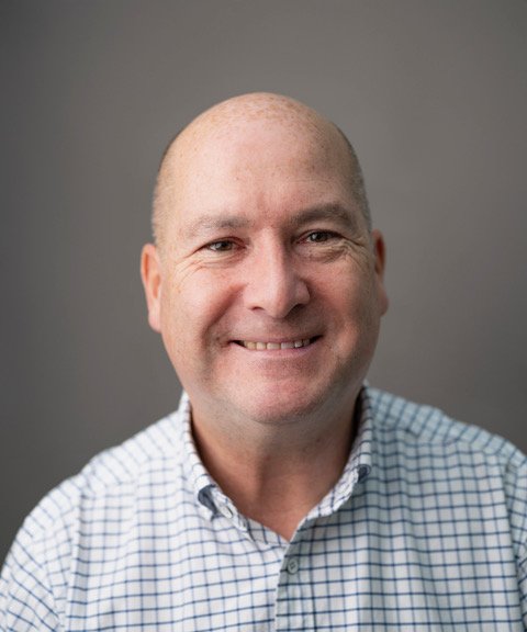 Close-up portrait of a bald man smiling, wearing a checkered shirt, against a gray background.