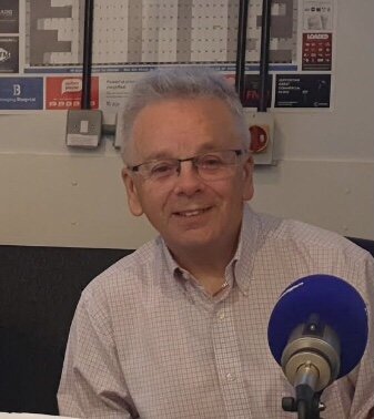 An older man with glasses and gray hair smiling in a radio studio, with a microphone in front of him.