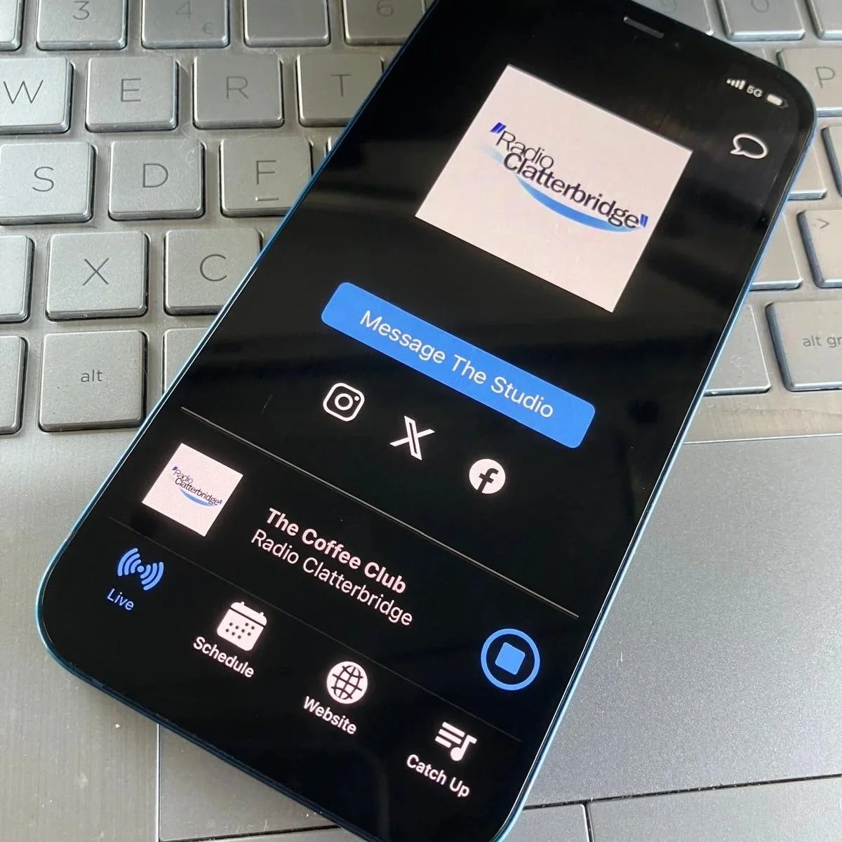 Smartphone displaying the logo and options for Radio Clatterbridge, a radio station, resting on a silver keyboard.