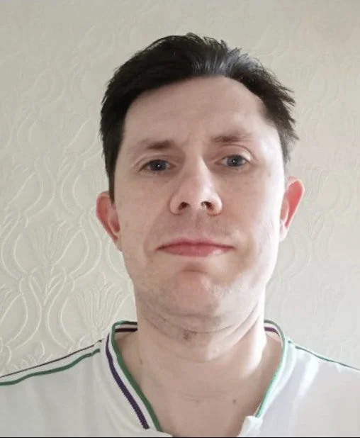 A young man with dark brown hair and fair skin taking a selfie indoors with a textured light-colored wall in the background.