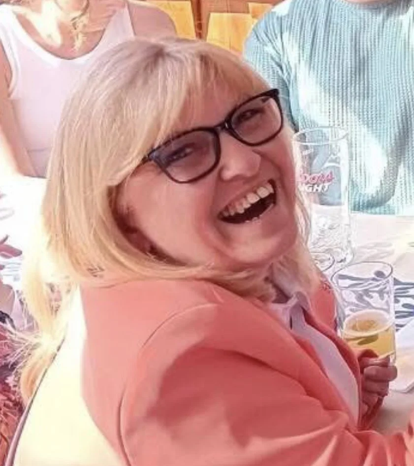A woman with blonde hair and glasses laughing joyfully at a social gathering, with drinking glasses and other people in the background.