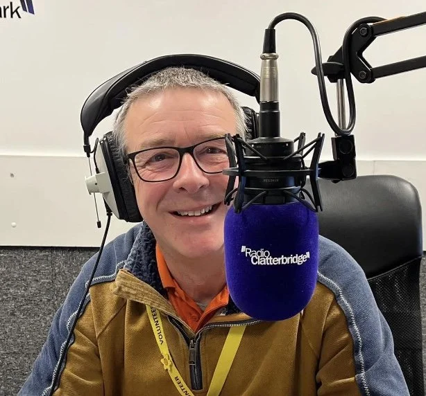 A man with glasses and gray hair wearing headphones, sitting in front of a microphone with a blue windscreen labeled '
 Radio Clatterbridge,' smiling.
