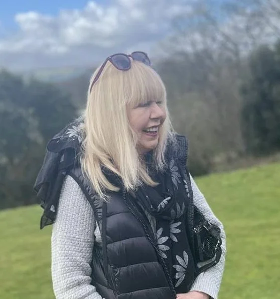 Woman with blonde hair, wearing sunglasses, a black vest, and a grey sweater, smiling outdoors on a grassy area with trees and cloudy sky in the background.