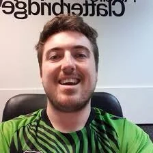 A young man with brown hair and a beard smiling, wearing a green and black patterned shirt, sitting in front of a wall with partially visible text.