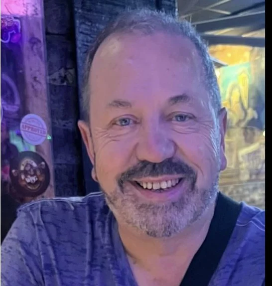 A smiling middle-aged man with a beard and short hair, wearing a dark shirt, in front of a colorful, neon-lit background.