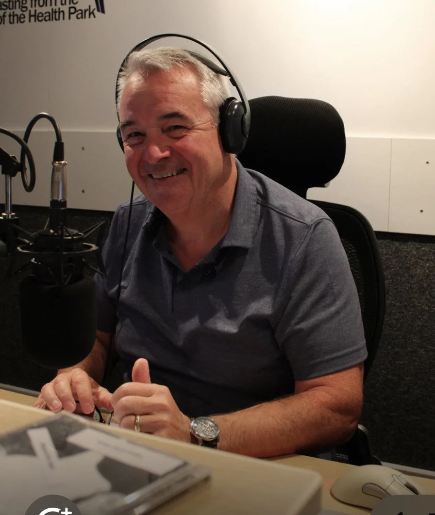 A middle-aged man with gray hair smiling and wearing headphones, sitting in front of a microphone in a recording studio or radio station.