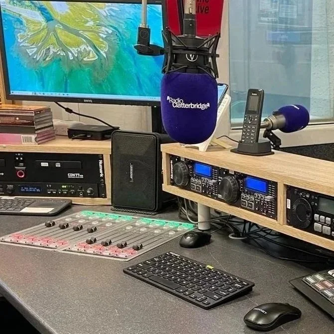 Radio broadcasting studio with computer monitors, audio equipment, microphones, and a mixing console on the desk.