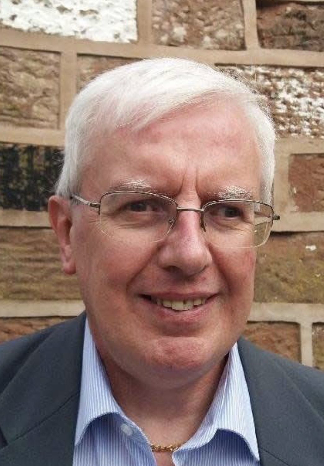 A smiling elderly man with white hair, glasses, and a dark suit jacket, wearing a light blue shirt, standing in front of a decorative stone wall.