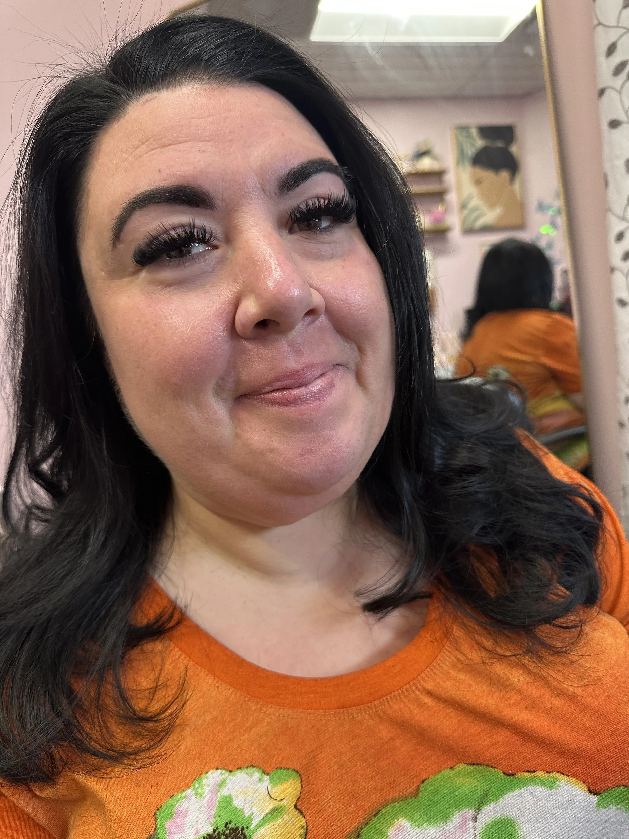 A woman with black hair and makeup, wearing an orange shirt with floral design, smiling in front of a mirror in a salon or beauty shop.