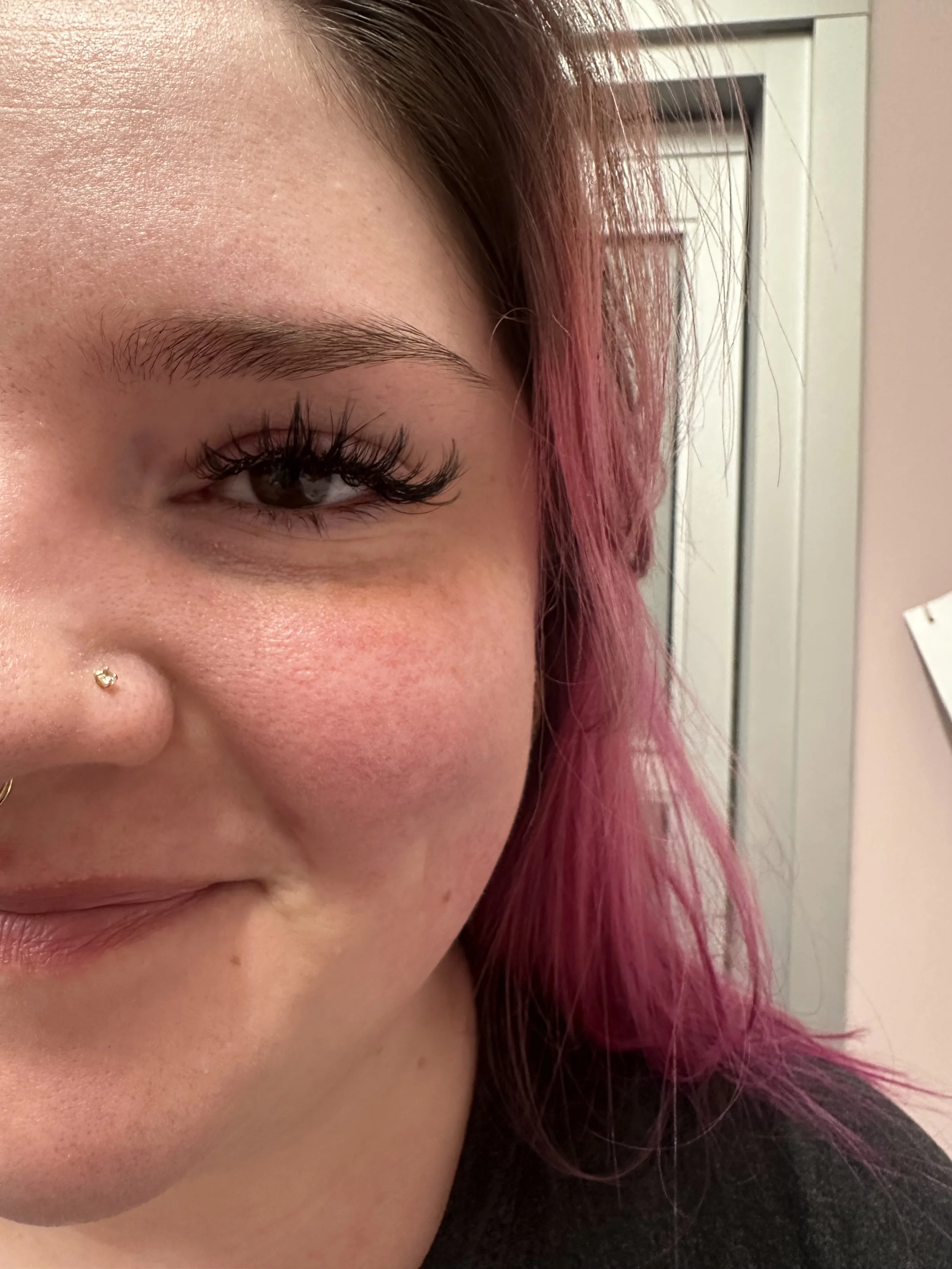Close-up of a woman's face showing her eye, eyebrow, and part of her nose with a nose piercing, with pink hair and a blurred background.